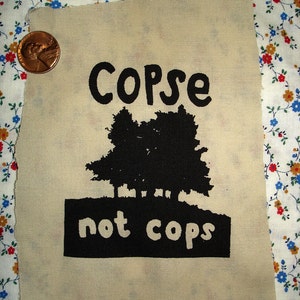 Könnte beinhalten: Ein beigefarbener Stoff-Patch mit schwarzem Text und einer schwarzen Silhouette von Bäumen. Der Text lautet "Copse not cops" und die Website "retirementfund.etsy.com" ist unten aufgedruckt.