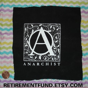 Può includere: Toppa in tessuto nero con una lettera "A" stilizzata bianca all'interno di un cerchio circondato da un motivo floreale decorativo. Il testo "ANARCHIST" è stampato sotto il disegno.
