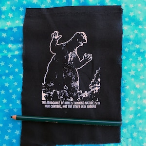 May include: Black and white patch with a silhouette of a monster, possibly Godzilla, standing in front of a building. The text reads "The arrogance of man is thinking nature is in our control, not the other way around."