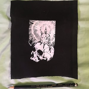 Puede incluir: Parche blanco y negro con una calavera y una vela en la parte superior. La vela tiene una llama y está rodeada de una luz radiante. El parche está hecho de tela y tiene una textura rugosa.