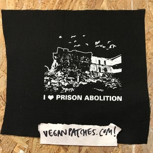 May include: Black and white iron-on patch with a graphic of a building in ruins and the text "I ♥ PRISON ABOLITION".