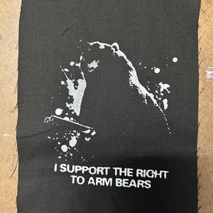 Könnte beinhalten: Schwarz-weißes Patch mit einer Silhouette eines Bären, der ein Gewehr hält. Der Text "I support the right to arm bears" ist unter dem Bären gedruckt.