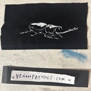 May include: A black fabric patch with a white beetle illustration. Beneath it, a black strip displays the text "VEGANPATCHES.COM" in white. The background is a textured, off-white surface with blue paint.