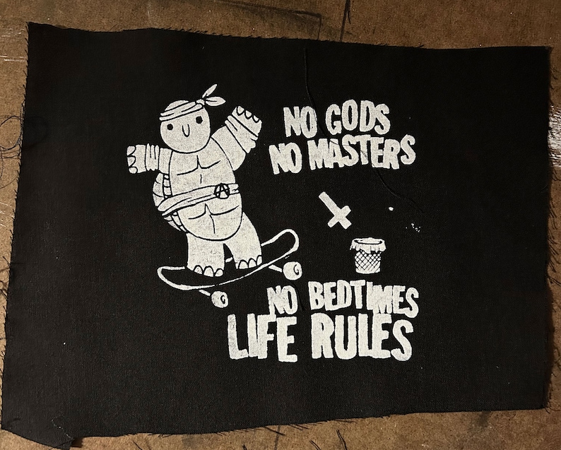 May include: Black and white patch with a cartoon turtle skateboarding. The text reads "No Gods No Masters No Bedtimes Life Rules".