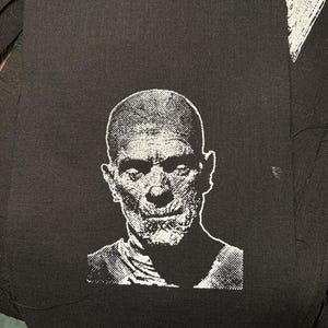 May include: Black and white embroidered patch of a man's face with a serious expression. The patch is on a black fabric background.