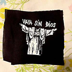 May include: Black fabric patch with white text that reads "VAYA SIN DIOS" and a white silhouette of a person with arms outstretched.