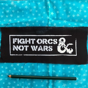 Könnte beinhalten: Ein schwarzer Aufnäher mit weißem Text "FIGHT ORCS NOT WARS" und einem Drachenlogo. Der Aufnäher befindet sich auf blauem Stoff mit weißen Sternen. Ein grüner Bleistift liegt darunter.