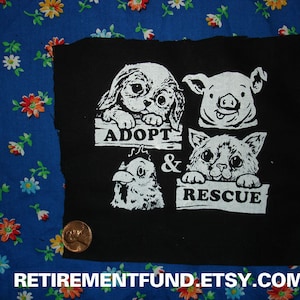 May include: Black fabric patch with white text and images of a dog, pig, cat, and chicken. The text reads "Adopt & Rescue".