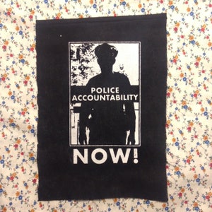 May include: Black and white patch with a silhouette of a police officer and the text "Police Accountability Now!"