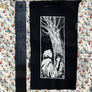 May include: Black and white embroidered patch featuring a hooded figure reaching towards a large tree with branches reaching up to a starry sky.