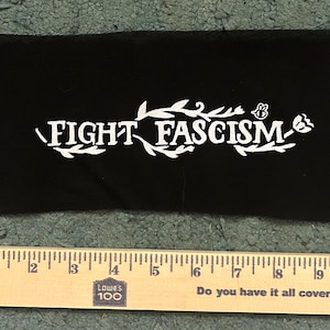 Op de afbeelding: Een rechthoekige zwarte stoffen patch met de witte opdruk FIGHT FASCISM, omringd door decoratieve illustraties van bladeren en een bij. Een houten liniaal ligt onder de patch en toont een lengte van ongeveer 23 cm.