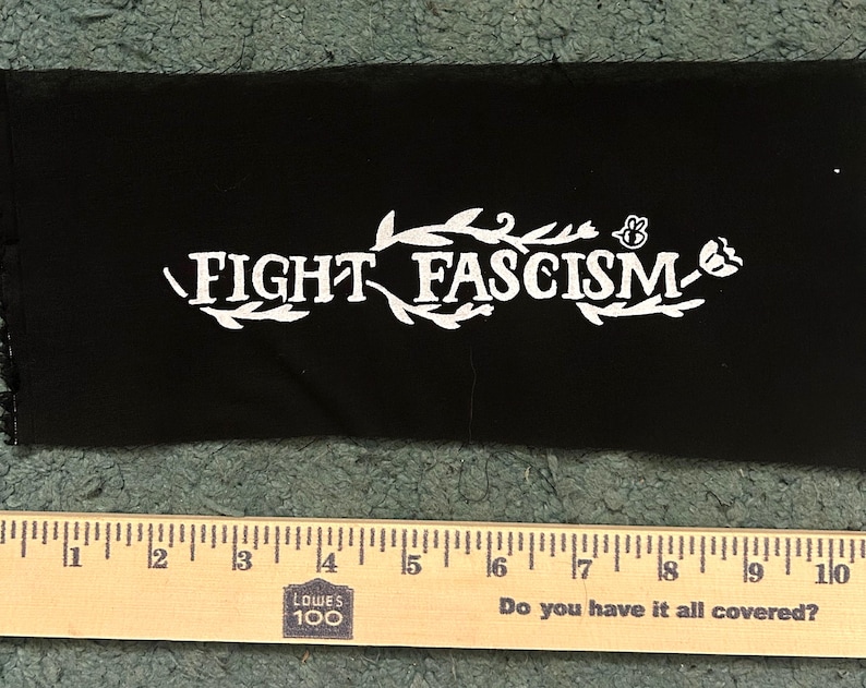 Op de afbeelding: Een rechthoekige zwarte stoffen patch met de witte opdruk FIGHT FASCISM, omringd door decoratieve illustraties van bladeren en een bij. Een houten liniaal ligt onder de patch en toont een lengte van ongeveer 23 cm.