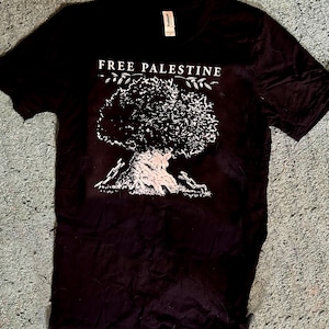 May include: Black t-shirt with the words "FREE PALESTINE" above a white graphic of a tree. The tree has a detailed trunk and branches with leaves. The shirt is made of a soft material.