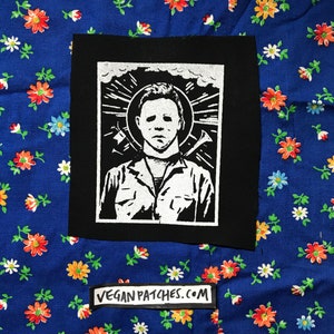 May include: Black and white iron-on patch featuring a stylised image of Michael Myers from the Halloween franchise. The image depicts him with a white mask and a halo, holding a butcher knife and a hook. The patch has the text "VEGAN PATCHES.COM" printed on it.