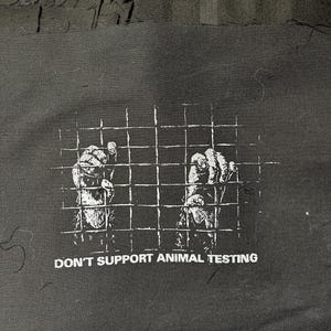 Puede incluir: Impresión gráfica en blanco y negro sobre un fondo de tela negro. El gráfico muestra dos figuras en una jaula con el texto "No apoyes las pruebas en animales".