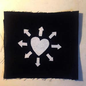May include: A black fabric patch with a white heart surrounded by eight white arrows pointing outwards.