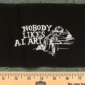 May include: A black fabric patch featuring white screen-printed text that reads NOBODY LIKES A.I. ART next to a vintage-style illustration of a person writing at a desk. A wooden ruler is positioned below, showing a measurement of 17 inches.