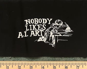 Puede incluir: Un parche de tela negra con texto serigrafiado en blanco que dice NOBODY LIKES A.I. ART junto a una ilustración vintage de una persona escribiendo en un escritorio. Una regla de madera se sitúa debajo, mostrando 43,18 cm.