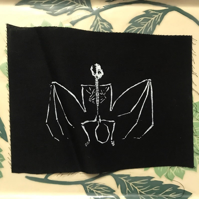 May include: A black fabric patch featuring a white skeletal bat design. The bat's wings are spread, and the detailed illustration includes the rib cage, spine, and skull. The patch has a raw edge.