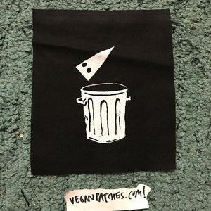 May include: Black fabric patch featuring a white graphic of a bin with a slice of pizza falling into it. The text "VEGAN PATCHES.COM!" is written on a white piece of paper below the patch.