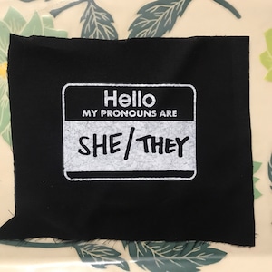 May include: A black fabric patch with a white name tag design. The name tag reads "Hello MY PRONOUNS ARE SHE/THEY" in a bold, sans-serif font. The patch is rectangular with slightly frayed edges.