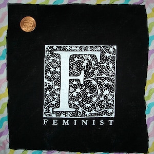 May include: Black fabric patch with a white stylized letter "F" surrounded by a floral design. The text "FEMINIST" is printed below the letter.