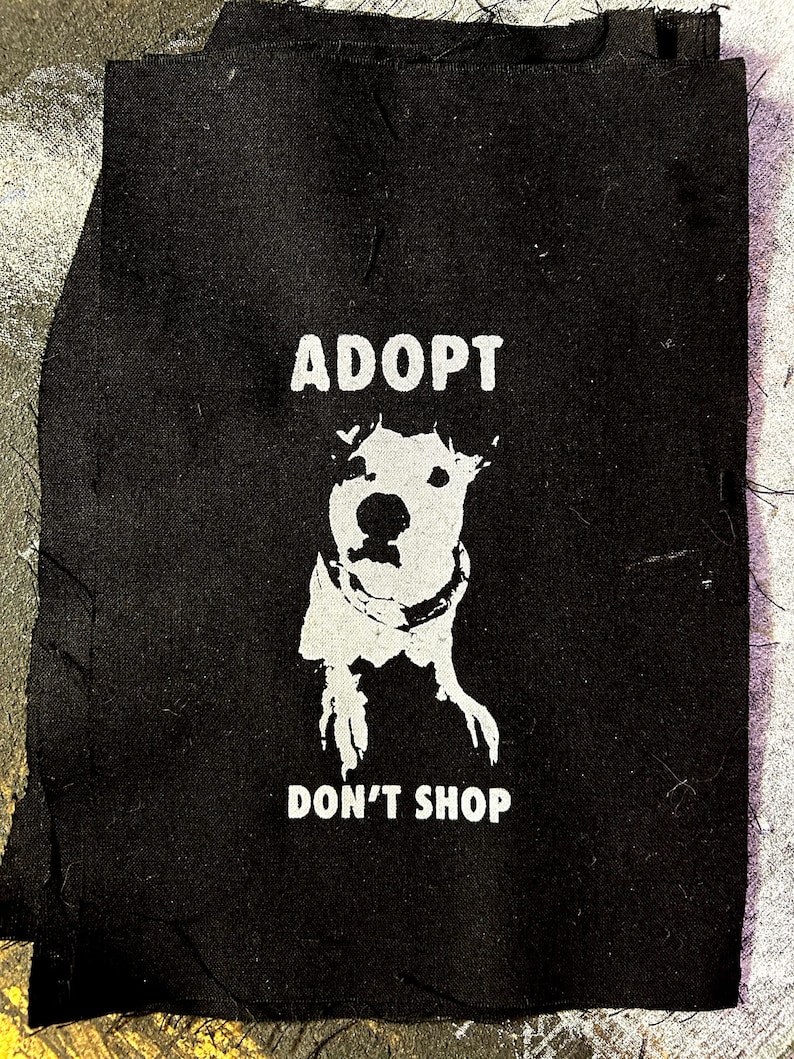 May include: Black fabric patch with white text that reads "Adopt Don't Shop" and a white silhouette of a dog's head.