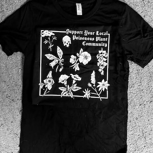 Może przedstawiać: Czarny t-shirt z białą grafiką przedstawiającą różne ilustracje roślin i napis "Support Your Local Poisonous Plant Community" w białej prostokątnej ramce.