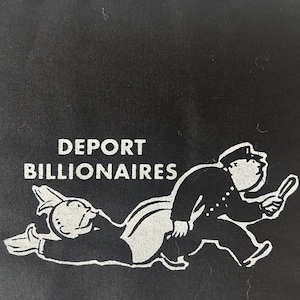 Op de afbeelding: Zwarte stof met de tekst "DEPORT BILLIONAIRES" boven een witte afbeelding van een politieagent die de Monopoly-man achtervolgt. De stof heeft een getextureerde uitstraling en een ruwe rand.