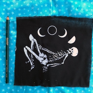 May include: Black fabric patch with a white skeleton design and a crescent moon pattern. The skeleton is lying on its back with its arms outstretched.