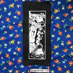 Puede incluir: Parche termoadhesivo en blanco y negro que representa a una mujer siendo arrastrada a un caldero por una criatura con cuernos. El parche tiene un fondo negro y un diseño blanco. El texto "veganpatches.com" es visible en la parte inferior de la imagen.