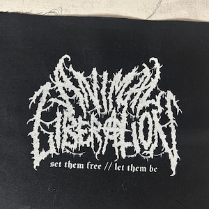 May include: Black fabric patch featuring white gothic lettering that spells "SINIC LIBERATION". The patch also includes the text "set them free // let them be" in a smaller font below the main text.