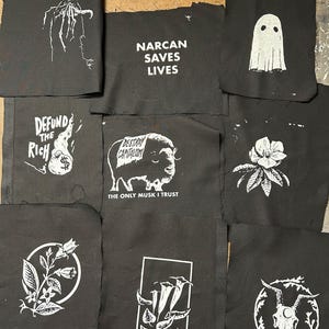 May include: A collection of black fabric patches with white graphic designs and text. Designs include a ghost, a bison with text, a floral design, a skull, and text that reads "NARCAN SAVES LIVES", "DEFUND THE RICH", and "DESTROY CAPITALISM".