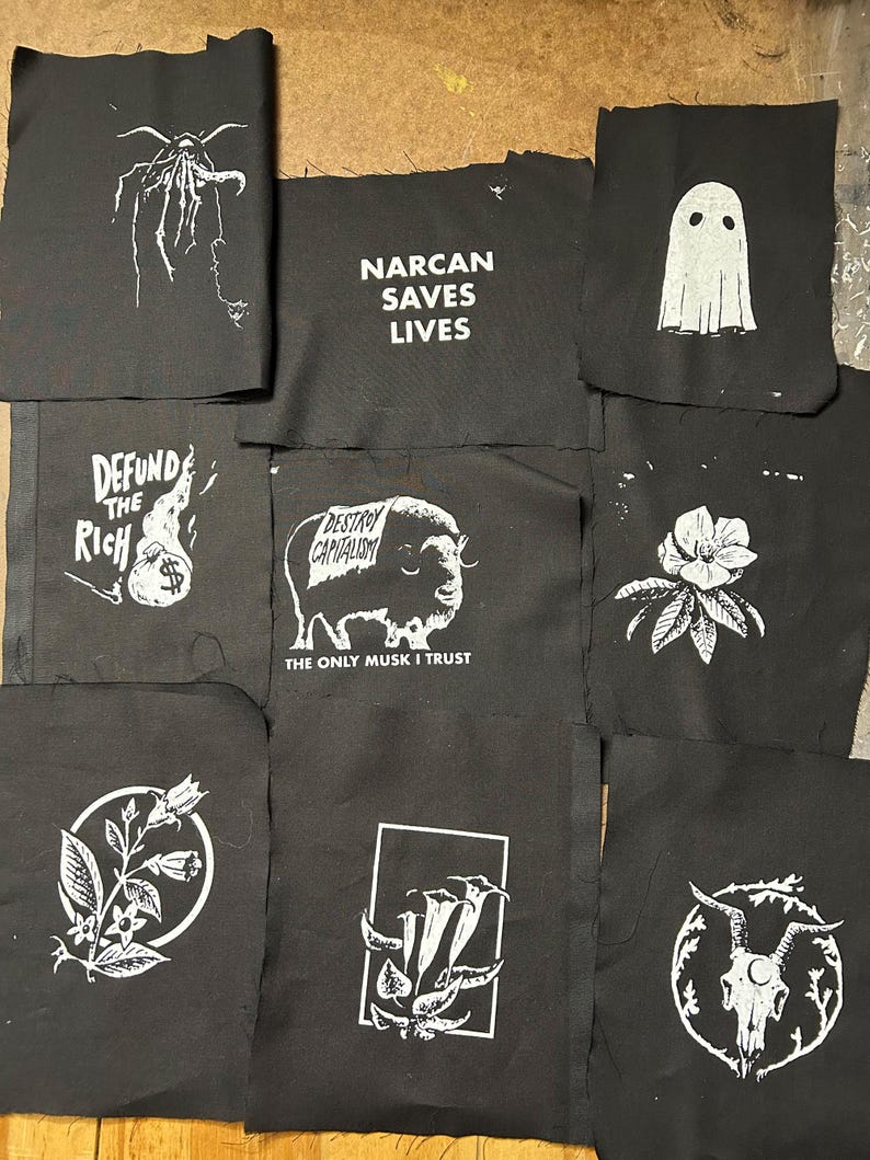 May include: A collection of black fabric patches with white graphic designs and text. Designs include a ghost, a bison with text, a floral design, a skull, and text that reads "NARCAN SAVES LIVES", "DEFUND THE RICH", and "DESTROY CAPITALISM".