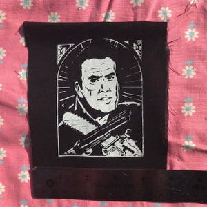 Könnte beinhalten: Schwarz-weißes Bügeleisen-Patch mit einem Porträt eines Mannes mit einer Kettensäge und einer Pistole. Der Mann trägt eine Lederjacke und hat einen ernsten Gesichtsausdruck.