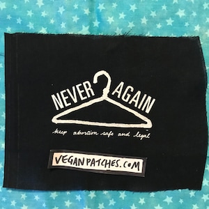 May include: Black fabric patch with white text that reads "NEVER AGAIN" with a white hanger graphic and the text "keep abortion safe and legal". The bottom of the patch has a smaller black fabric patch with white text that reads "VEGAN PATCHES.COM".