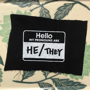 May include: Black fabric patch with a white name tag that reads "Hello, My Pronouns Are He/They".