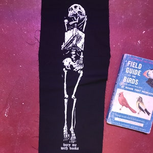 May include: A black fabric patch featuring a white skeleton illustration holding a book. The skeleton is wearing a crown and the text "bury me with books" is printed below the skeleton.