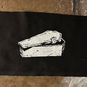 Könnte beinhalten: Ein schwarzer Stoff-Patch mit einer weißen Umrandung eines Holzsargs, dessen Deckel leicht geöffnet ist. Der Sarg ist in einem einfachen, cartoonartigen Stil gezeichnet.