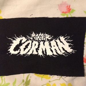 May include: Black fabric patch with white embroidered text that reads "Roger Corman" in a stylized, jagged font.