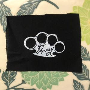 May include: Black fabric patch with a white outline of a knuckle duster and the text "go away" in a cursive font.