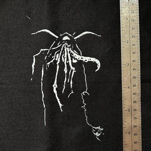 Może przedstawiać: Czarny materiałowy naszywka z białym nadrukiem stylizowanego stworzenia z mackami. Stworzenie ma około 15 cm wysokości.