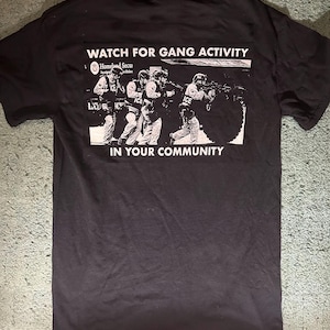 May include: A black short-sleeved t-shirt featuring a white graphic print on the back. The print depicts law enforcement officers in tactical gear with the text WATCH FOR GANG ACTIVITY above and IN YOUR COMMUNITY below the image.