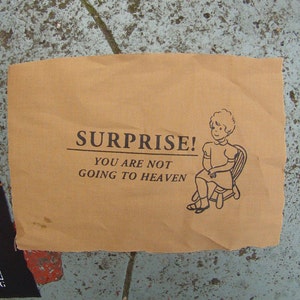 Puede incluir: Un cartel de papel marrón con el texto "SURPRISE! YOU ARE NOT GOING TO HEAVEN" y una ilustración en blanco y negro de una niña sentada en una silla.
