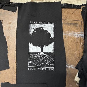 May include: Black fabric patch with white text and graphic. The text reads "TAKE NOTHING" above a tree illustration and "LEAVE EVERYTHING" below. The tree has exposed roots and a skeleton. Surrounding the patch are pieces of black and beige fabric.