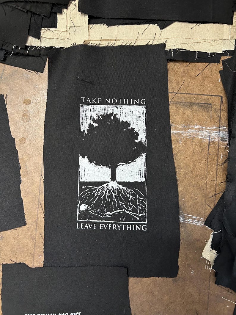 May include: Black fabric patch with white text and graphic. The text reads "TAKE NOTHING" above a tree illustration and "LEAVE EVERYTHING" below. The tree has exposed roots and a skeleton. Surrounding the patch are pieces of black and beige fabric.