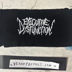 May include: Black rectangular patch with the words "EXECUTIVE DYSFUNCTION" in white, stylized, gothic lettering. Below the patch is a strip of paper with the text "VEGANPATCHES.COM". The background is a light, textured surface.