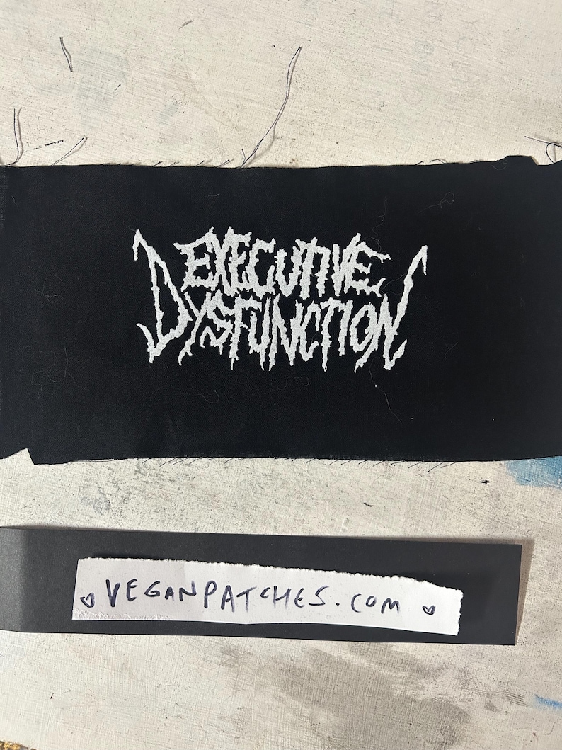 May include: Black rectangular patch with the words "EXECUTIVE DYSFUNCTION" in white, stylized, gothic lettering. Below the patch is a strip of paper with the text "VEGANPATCHES.COM". The background is a light, textured surface.