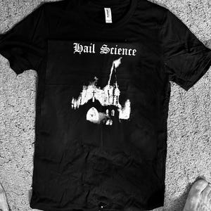 May include: Black t-shirt with the text "Hail Science" in gothic font above a white graphic of a church. The church graphic has a gothic architectural style. The t-shirt is laying flat on a gray surface.
