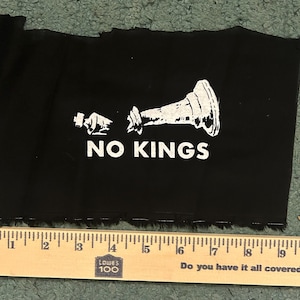 May include: A black rectangular fabric patch featuring a white screen-printed graphic of a broken chess king piece and the text NO KINGS. A wooden ruler is positioned along the bottom edge, showing the patch width is approximately 8 inches.
